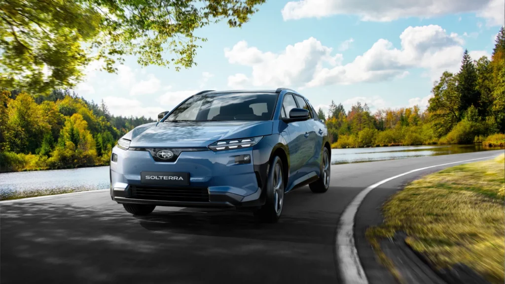 En blå Subaru Solterra SUV på vei forbi en innsjø og trær.