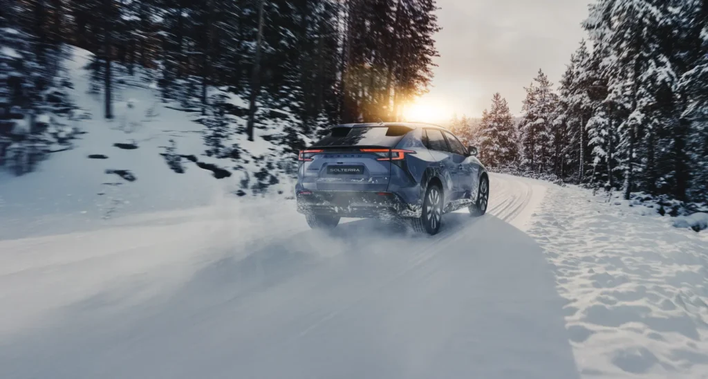 Blå Subaru Solterra SUV kjører på snødekt vei med trær.